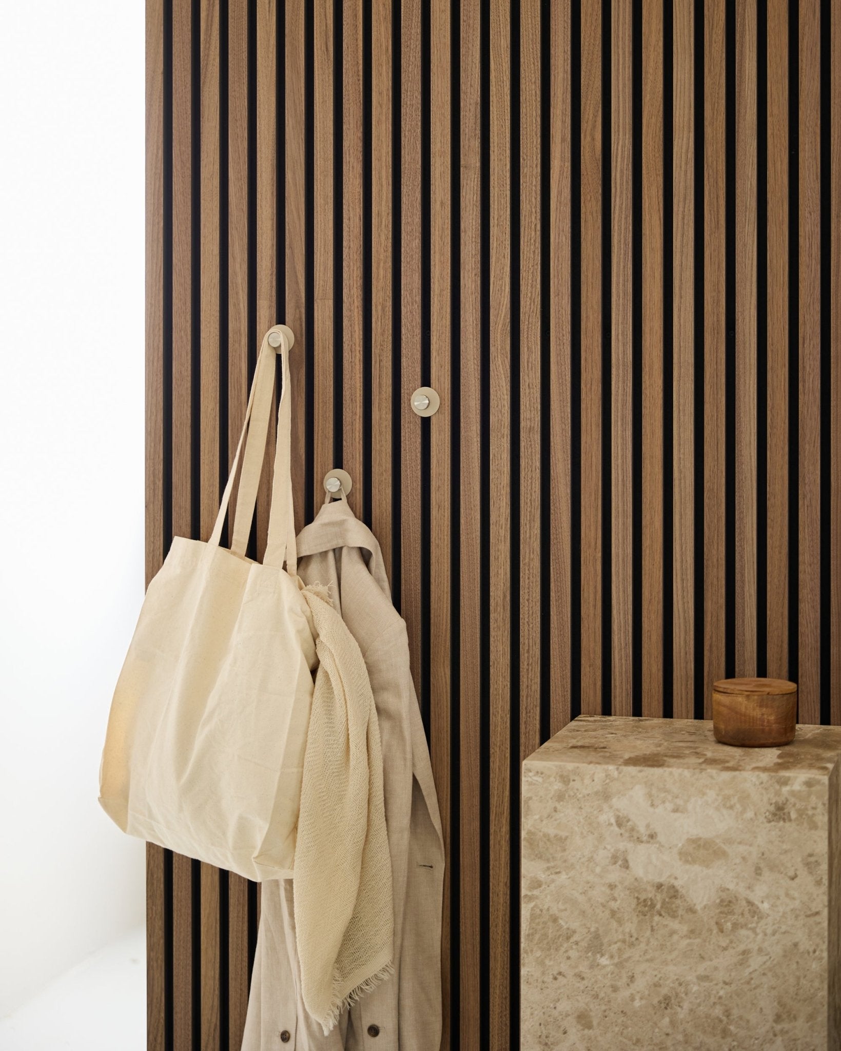 Un sac cabas beige et un tissu léger sont suspendus à une patère WoodUpp Norr (Beige/acier inoxydable Steel), fixée sur un vertical Akupanel Panneau acoustique à côté d’une surface en pierre.