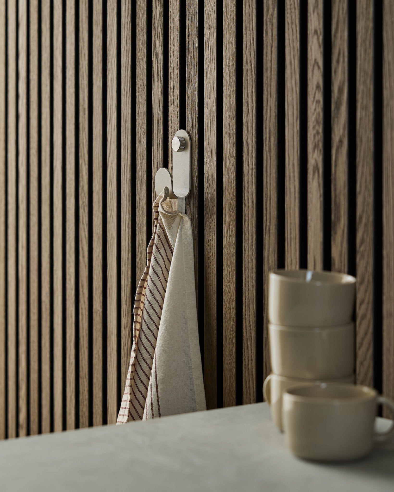 Une serviette rayée est suspendue à un WoodUpp Norr Single Hanger en beige, fixé sur un panneau acoustique à lattes de style nordique ; trois mugs beige sont empilés devant.
