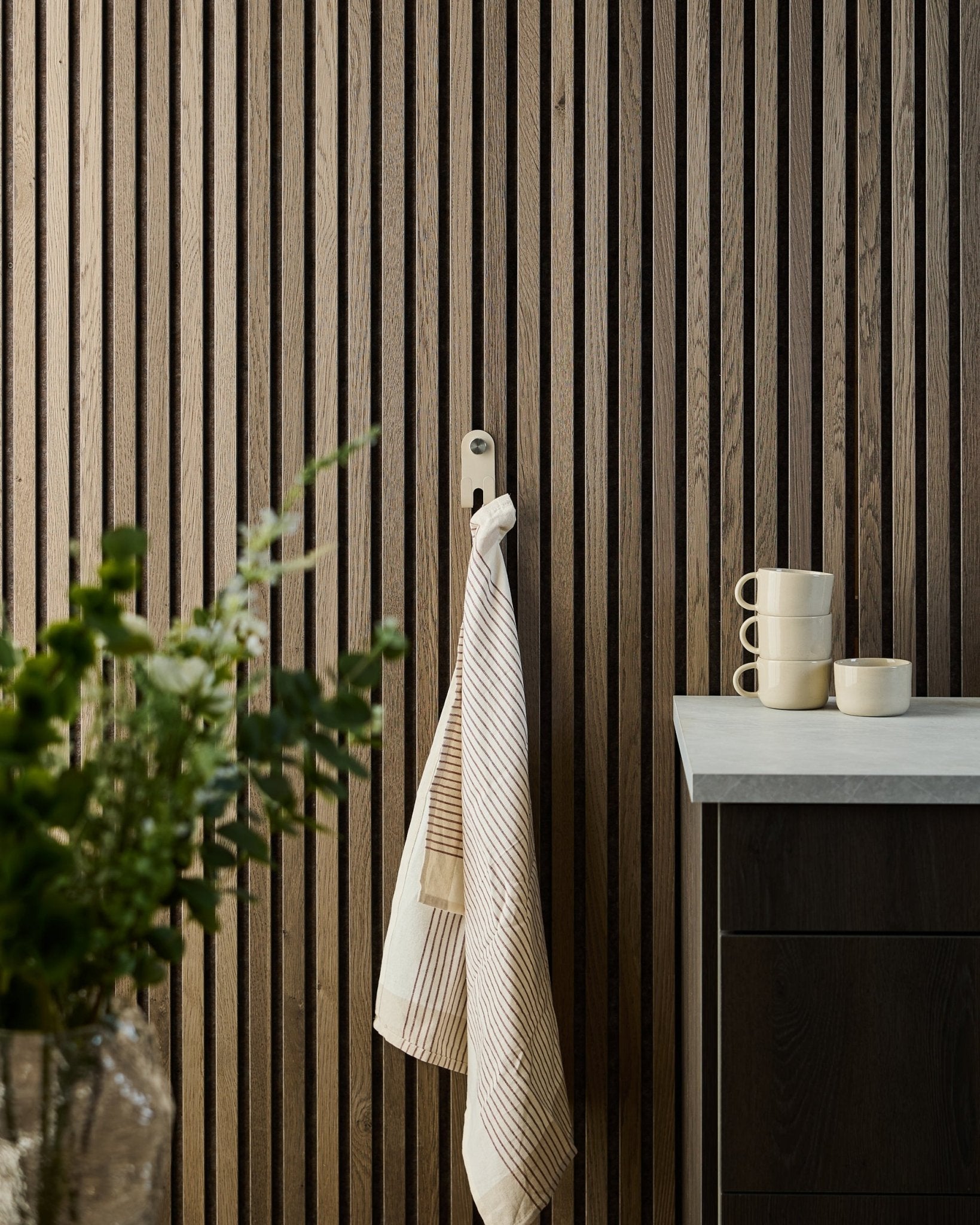 Un torchon rayé est suspendu au Norr Double Hanger de WoodUpp en beige, monté sur un panneau acoustique à lattes nordique ; des mugs, un bol et une plante verte sont disposés en dessous.