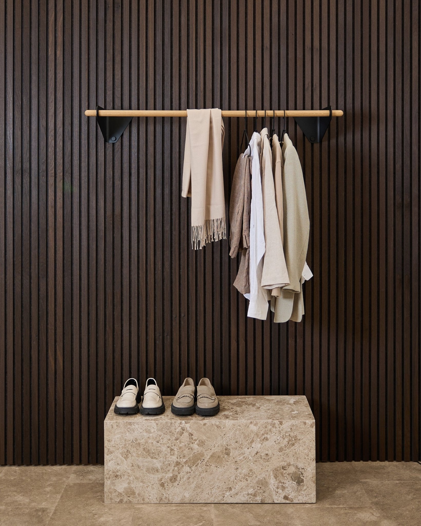Des vêtements aux tons neutres et une écharpe sont suspendus à une black de 115,9 cm Linn | Coat Rail par WoodUpp, au-dessus d’un banc en marbre, devant un panneau acoustique à lattes Akupanel foncé.