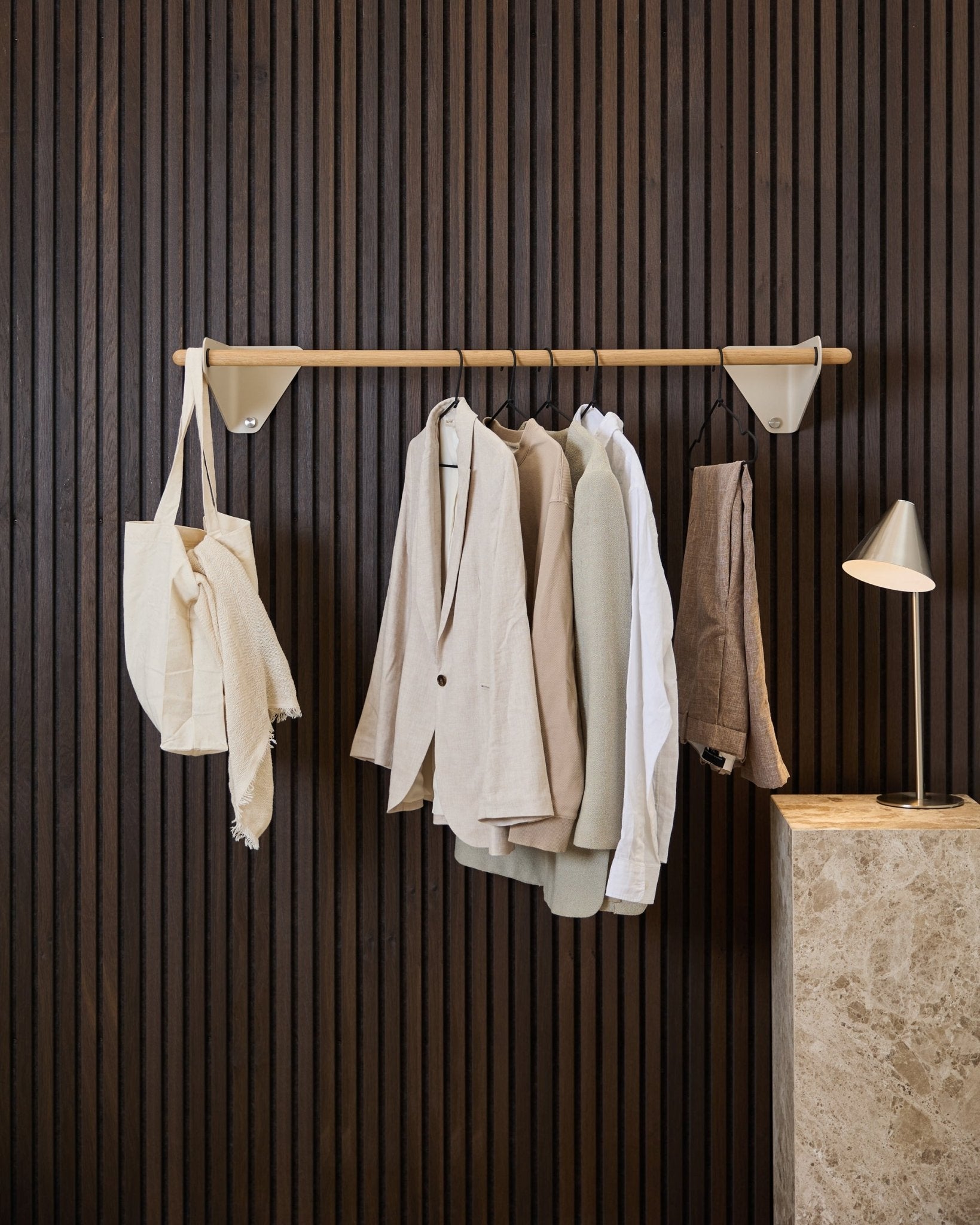 Un portemanteau WoodUpp Linn beige (115,9 cm) accueille des vestes, une chemise blanche et un tote bag crème devant un Akupanel Panneau acoustique en bois foncé et une lampe en marbre.