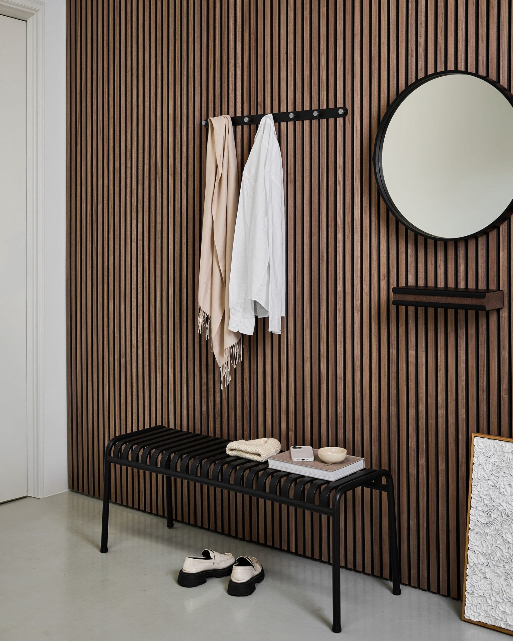 Entrée moderne avec un mur en panneau acoustique en bois à lattes nordique, un miroir rond, un banc black, le porte-manteau à patères WoodUpp Norr, et des chaussons sur un sol clair.