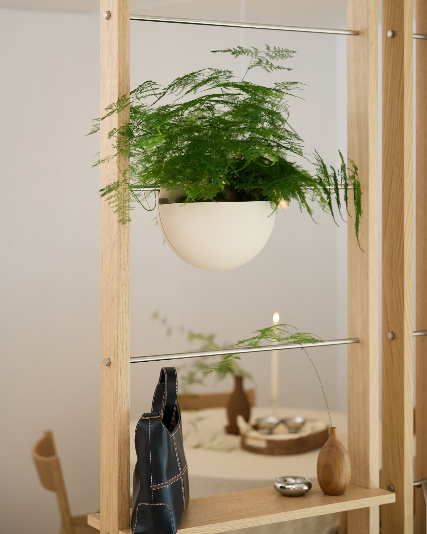 Une fougère verte, placée dans une jardinière WoodUpp Gro Soft Pocket BM2 de couleur beige clair, repose sur une étagère en bois au-dessus dun sac noir, dun vase et dune table à manger dressée.