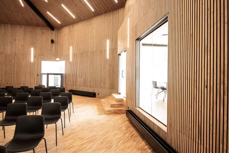 Salle de conférence spacieuse avec des panneaux acoustiques en bois Akupanel sur les murs et le plafond, des rangées de chaises noires, de grandes fenêtres et des luminaires verticaux.