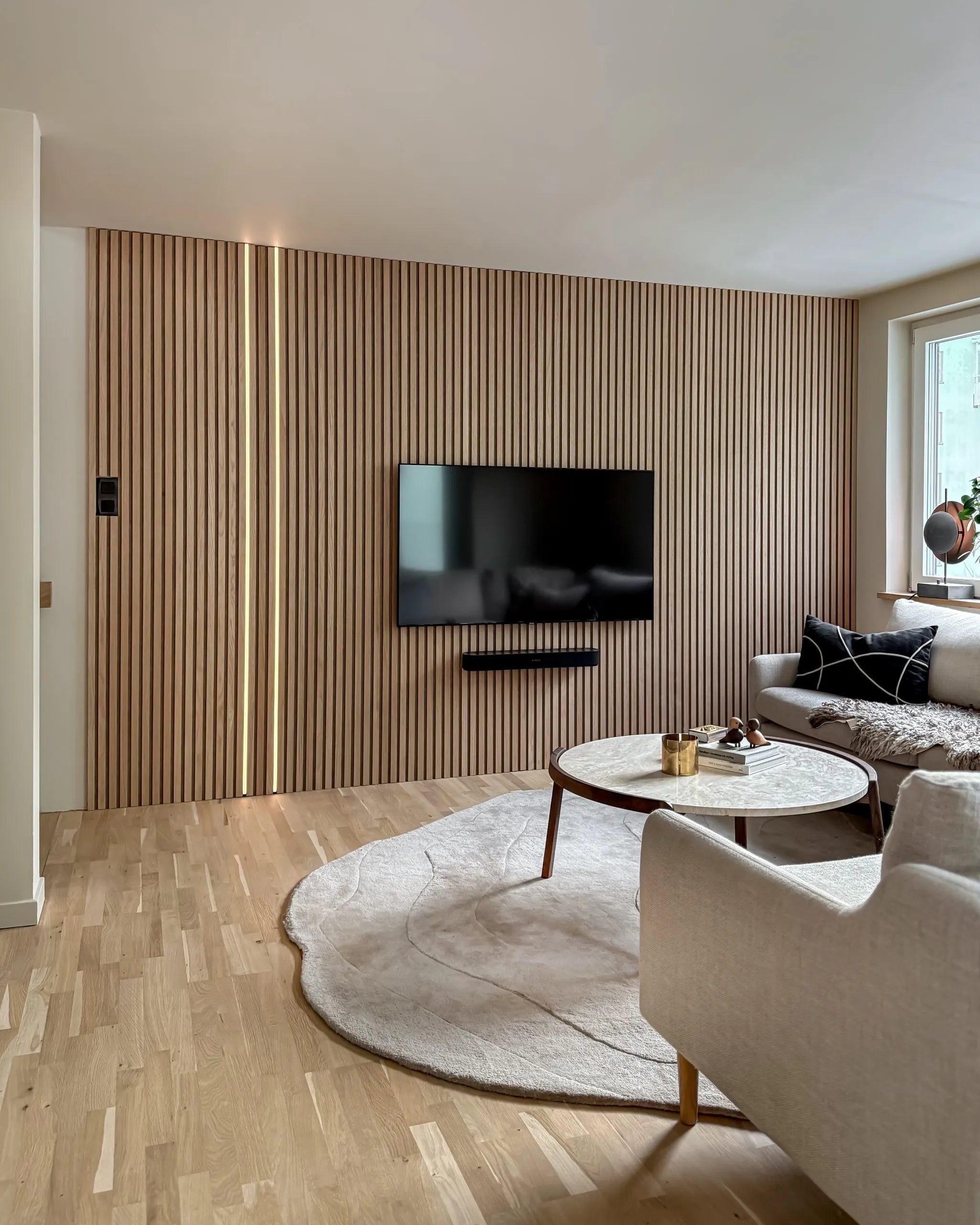 Salon moderne avec un mur daccent en lattes de bois acoustique Akupanel, une télévision montée, une table basse ronde, un canapé crème, un grand tapis sur un sol en bois clair.