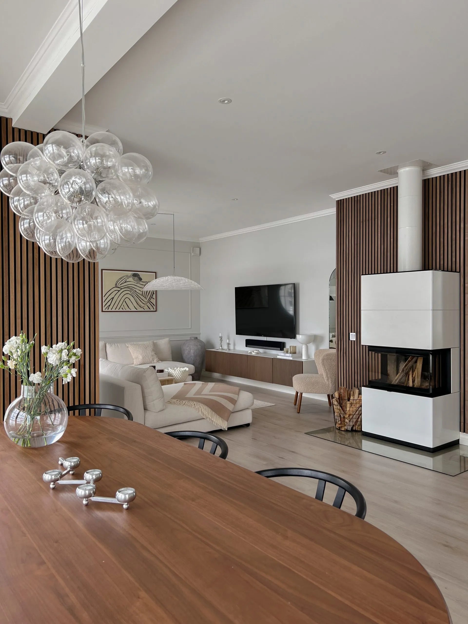 Salon et salle à manger modernes avec panneaux acoustiques en bois nordique, sièges confortables, télévision, cheminée élégante avec bois de chauffage empilé et lustre à bulles de verre.