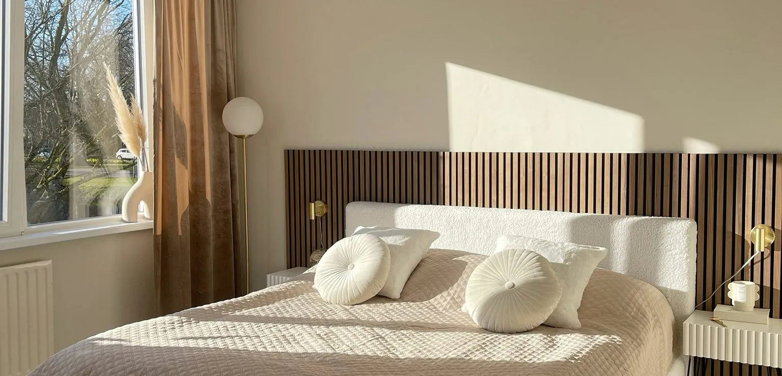 Chambre à coucher moderne ensoleillée avec un lit matelassé beige, des oreillers ronds, une tête de lit en lattes de bois Akupanel et un décor minimal à côté dune grande fenêtre avec des rideaux diaphanes.