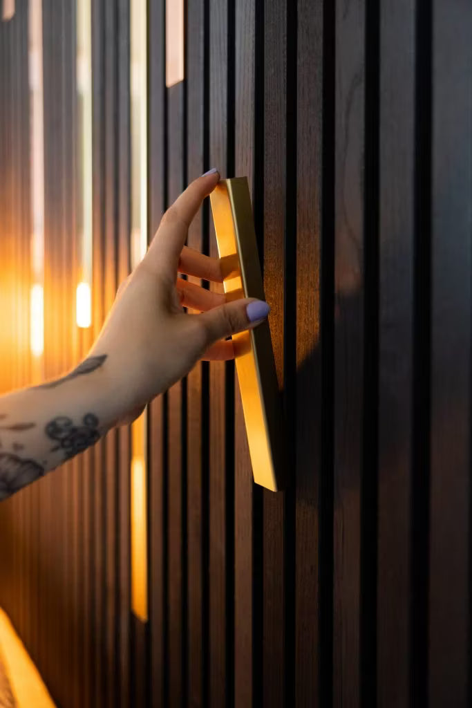 Une main tatouée aux ongles violet clair saisit une poignée dorée sur un mur en panneaux de bois acoustique Akupanel, sombre et épuré, mis en valeur par un éclairage chaleureux.