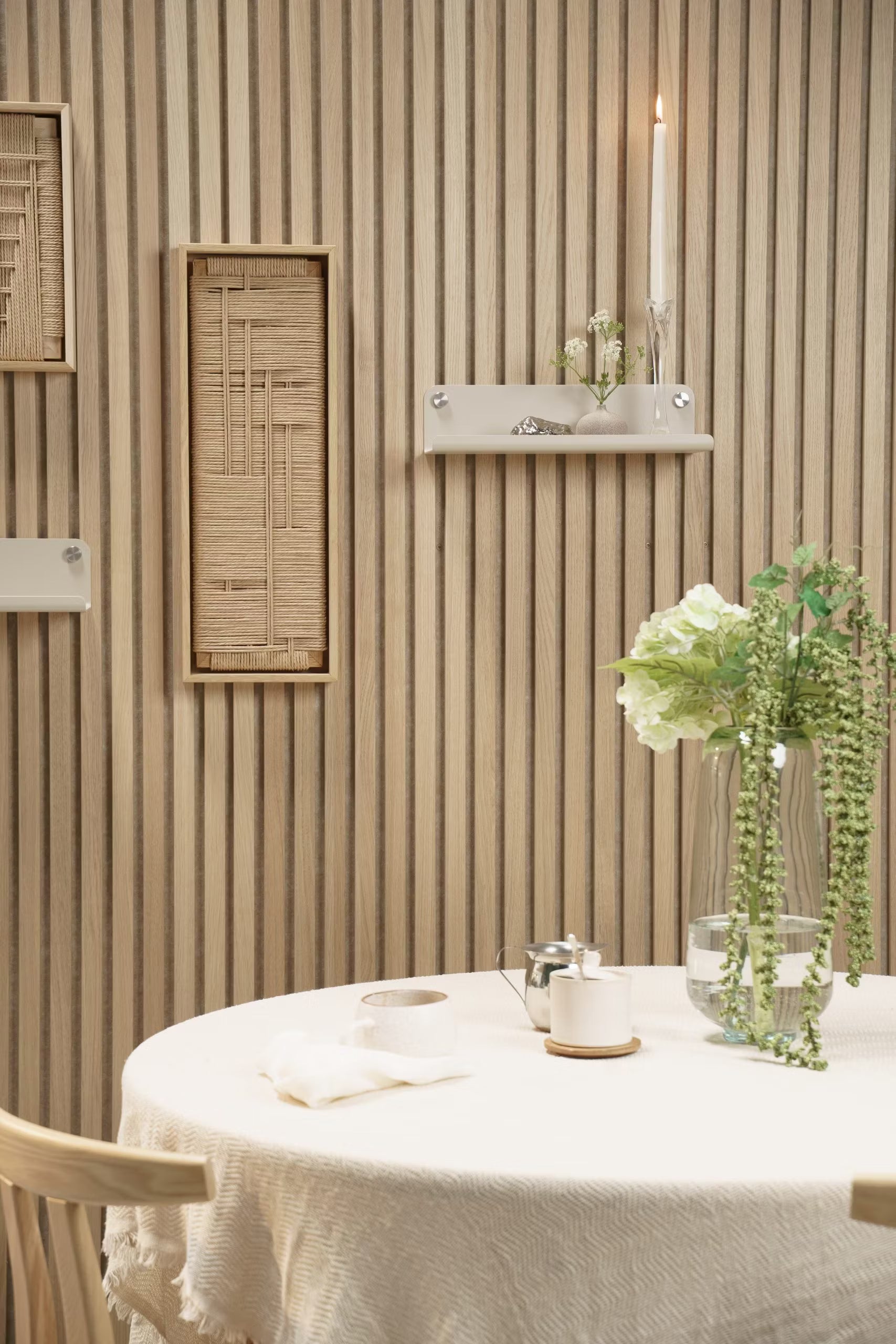 Une table ronde avec une nappe légère, des fleurs fraîches dans un vase et une bougie se trouve devant un mur de lattes en bois acoustique Akupanel de style nordique avec des étagères minimales.