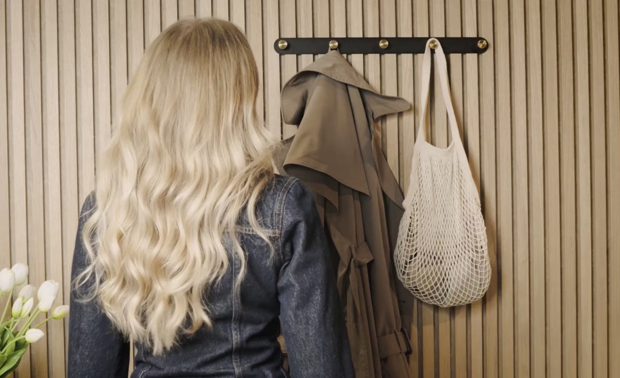 Une femme aux longs cheveux blonds se tient face à un porte-manteau contenant un trench-coat, un sac en filet blanc et des tulipes blanches fraîches, à côté dun panneau de bois acoustique Akupanel dans langle.