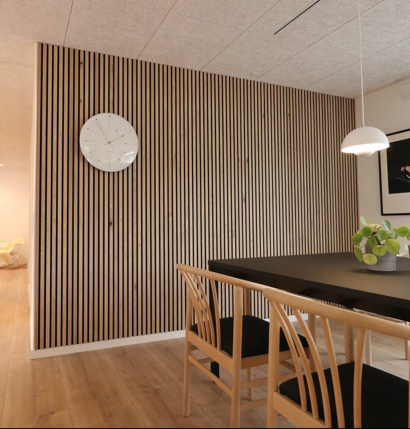 Salle à manger moderne avec un mur daccent en lattes de bois acoustique Akupanel nordique, une table noire, des chaises en bois avec des sièges noirs et un centre de table composé de plantes en pot.