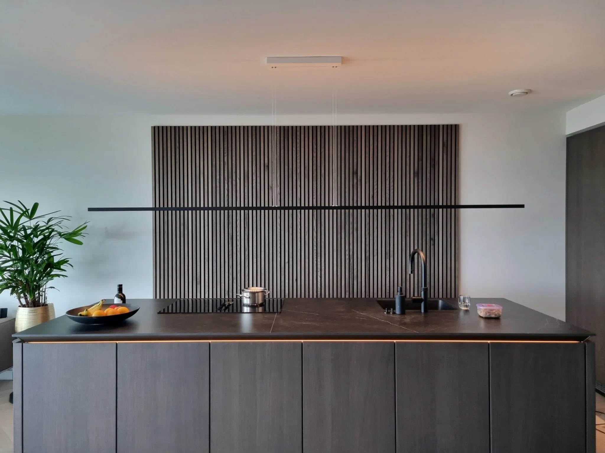 Cuisine moderne avec un îlot sombre, un évier et une cuisinière encastrés, une coupe de fruits et une plante en pot. Des panneaux verticaux en bois acoustique Akupanel ornent le mur derrière lîlot.