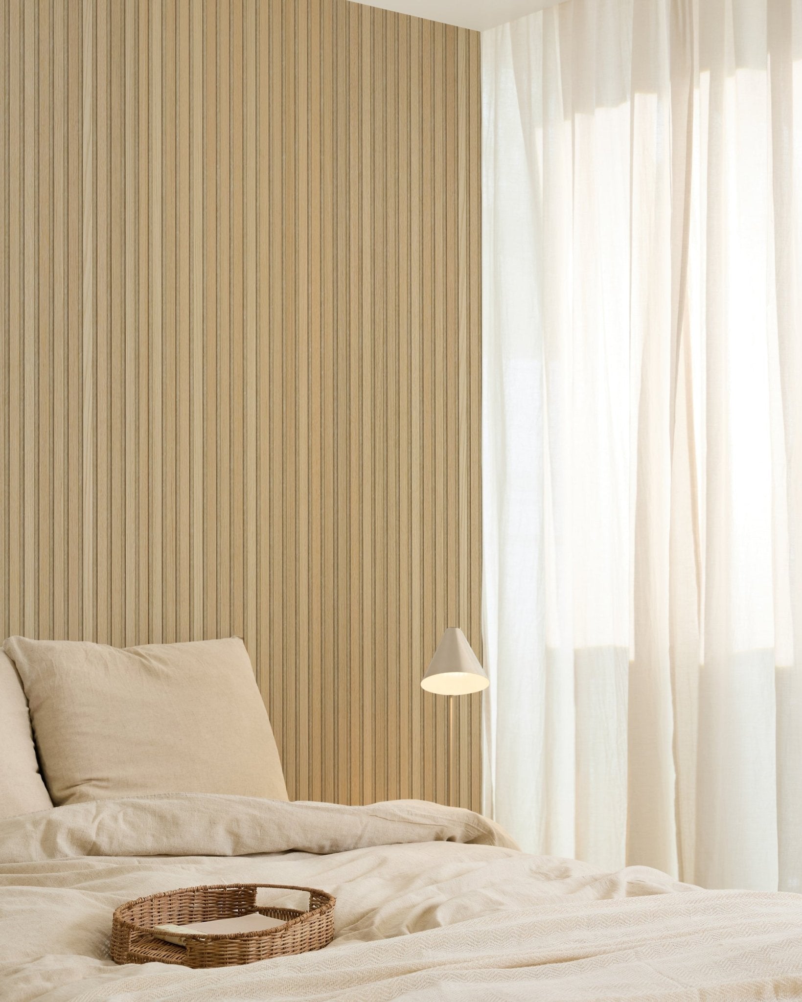 Chambre minimaliste avec du linge de lit beige, un plateau en osier, une applique murale moderne, un mur d’accent en panneau acoustique en bois Akupanel, et des rideaux voilages qui diffusent une douce lumière naturelle.