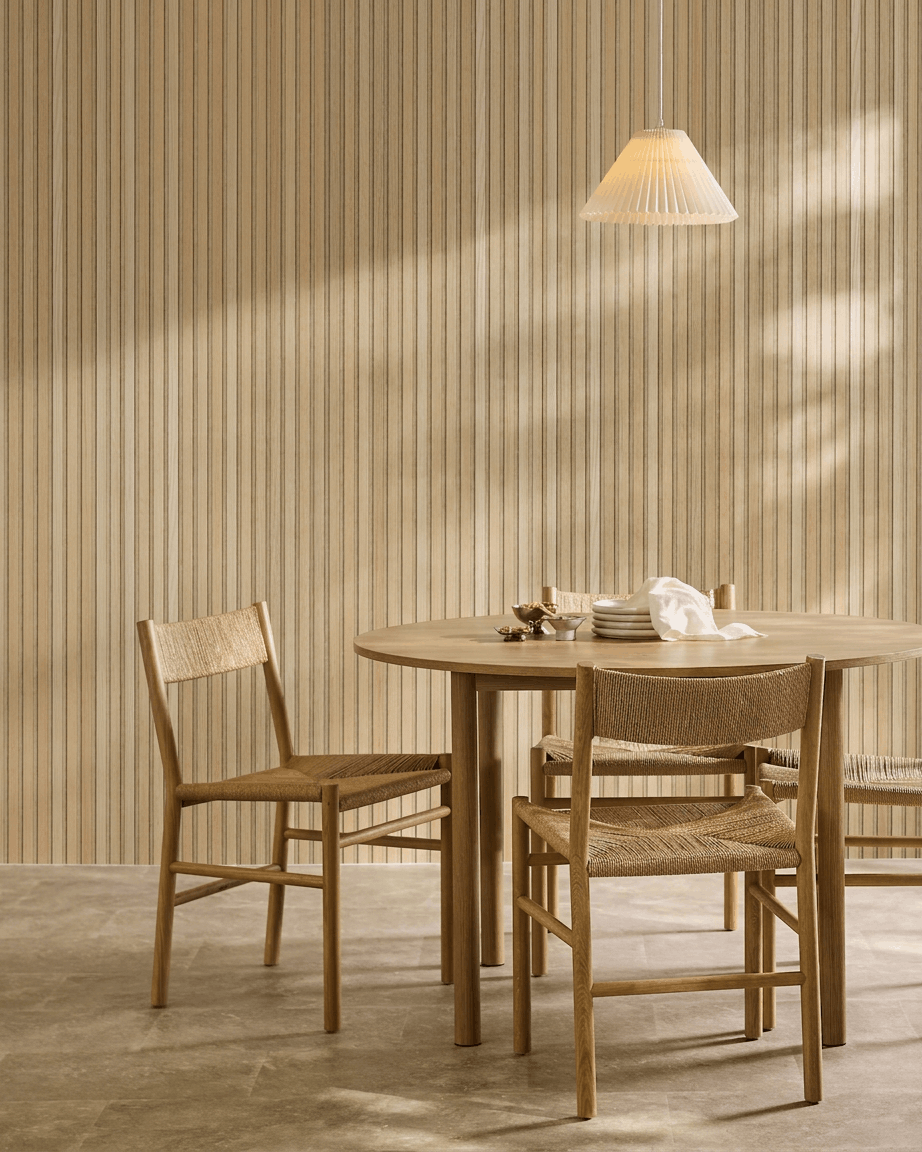 Salle à manger minimaliste avec une table ronde, des chaises tressées et une suspension, à côté d’un panneau acoustique en bois WoodUpp Akupanel en chêne classique avec feutre beige.