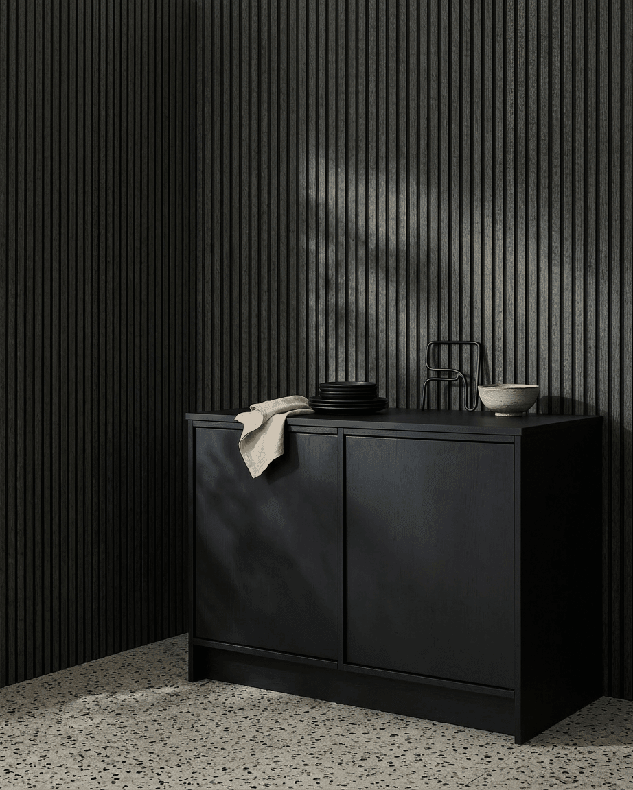 Un meuble black à côté d’un panneau acoustique en bois WoodUpp Akupanel en Charcoal/Black Felt, surmonté de bols et d’assiettes, sur un sol en terrazzo clair.