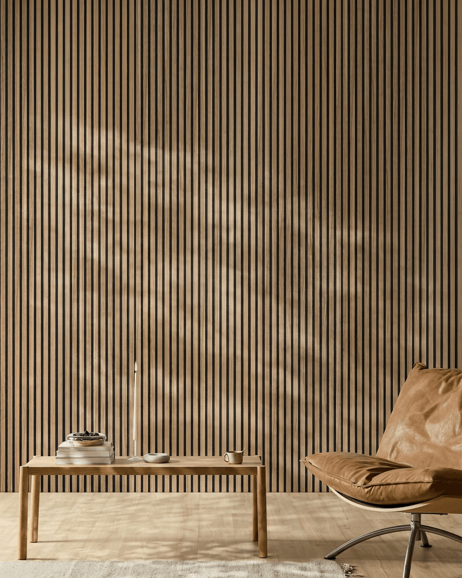 Pièce minimaliste avec un WoodUpp Akupanel Panneau acoustique (lattes en noyer anglais, feutre black), un fauteuil en cuir camel, une table en bois clair, une bougie, des livres, une tasse et un tapis blanc au soleil.