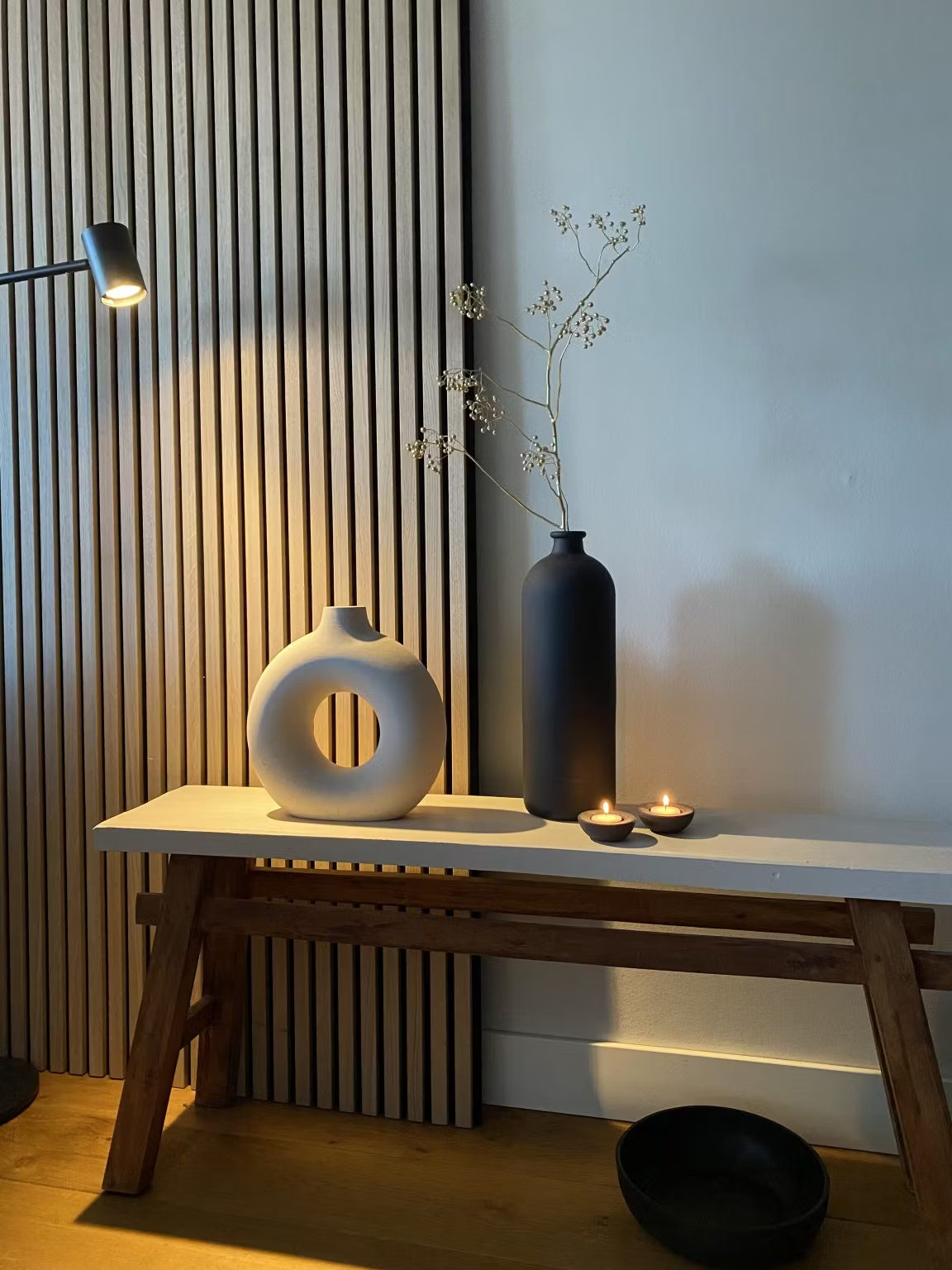 Décor minimaliste composé dun vase circulaire blanc, dun grand vase noir avec des tiges séchées, de deux bougies chauffe-plat et dun bol noir sur fond de panneau acoustique en lattes de bois Akupanel.
