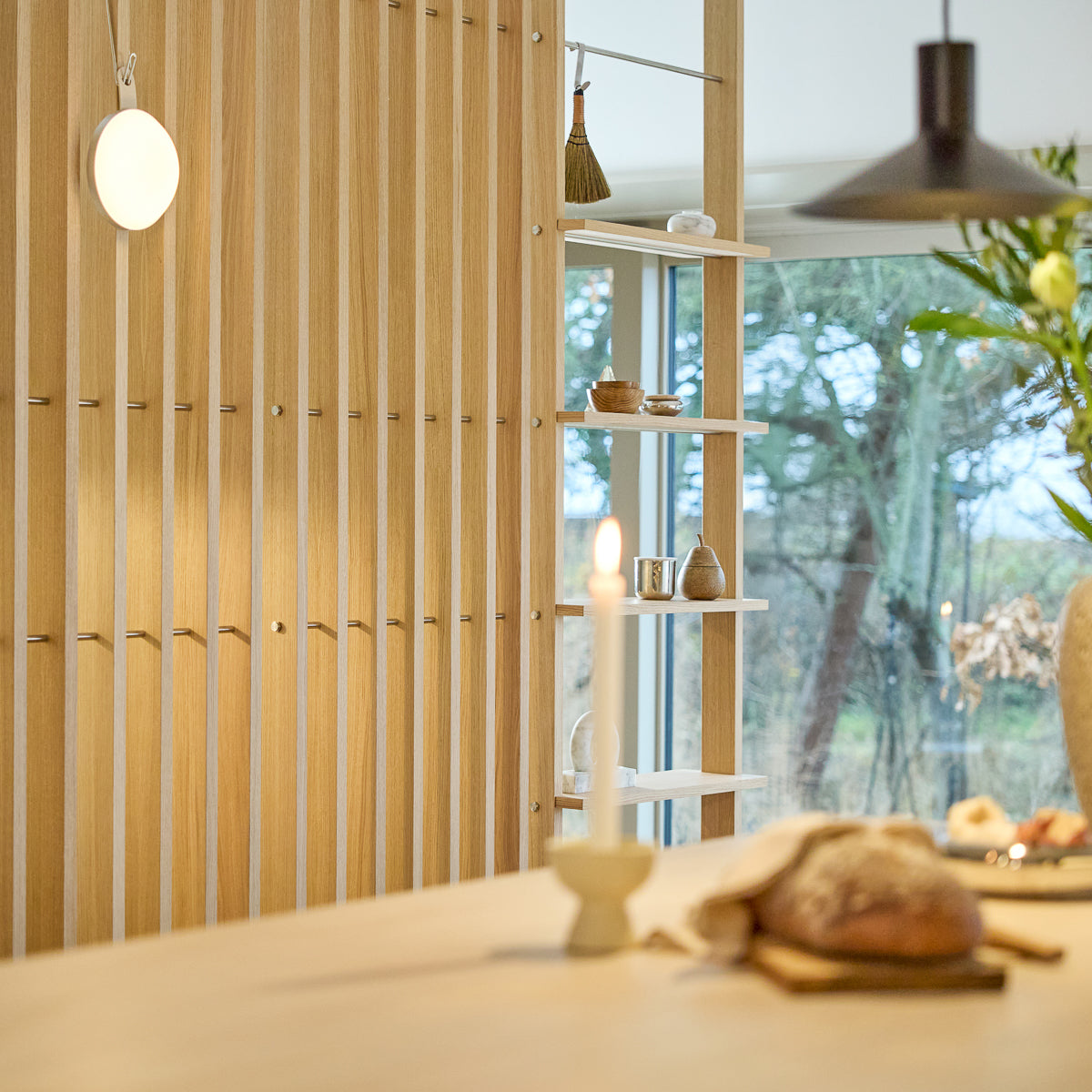 Intérieur minimaliste avec un paravent en bois Nordic EMBRACE, bougie allumée, pain frais sur la table, décor sélectionné sur les étagères et fenêtre encadrant des arbres à l'extérieur.