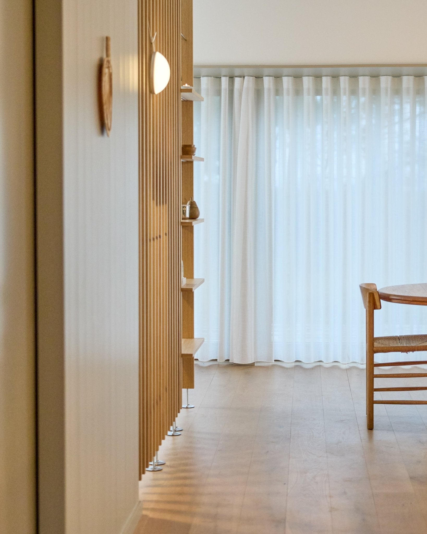Pièce minimaliste avec une cloison séparatrice WoodUpp Embrace du sol au plafond, un sol en bois, des étagères, une chaise et des rideaux voilages devant des fenêtres large.