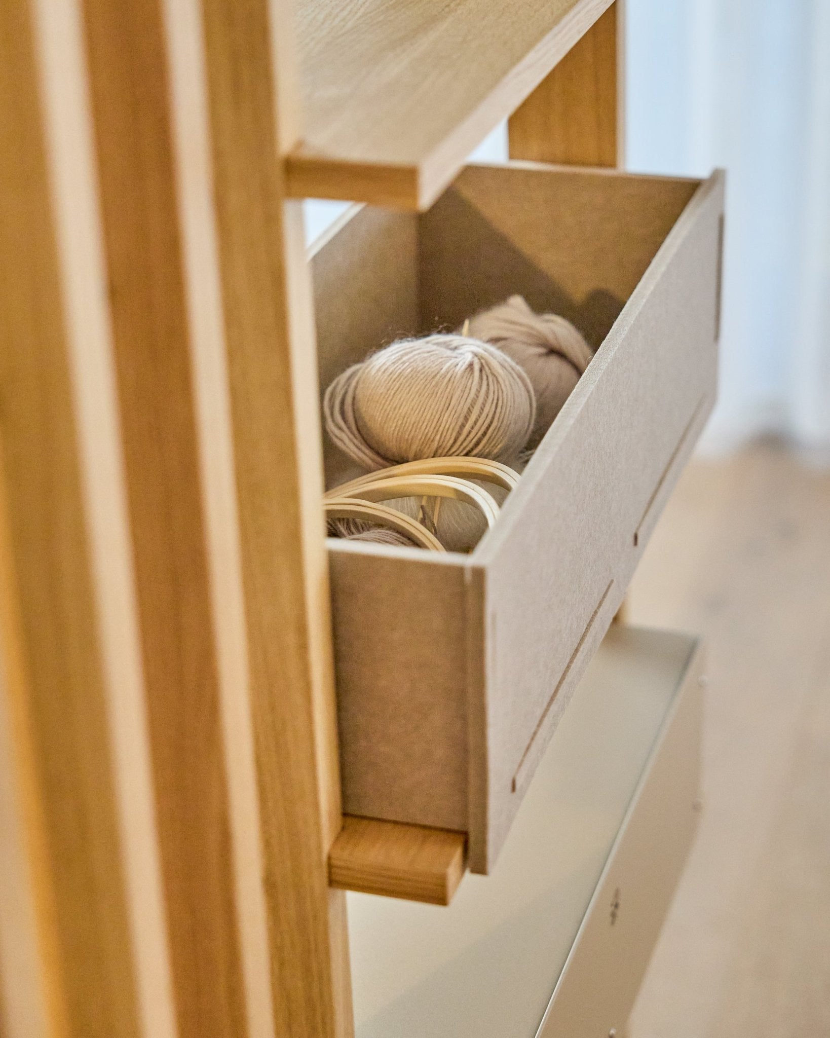 La WoodUpp Vigg Felt Box / BM2 en beige accueille des pelotes de laine légères et des tambours à broder en bois sur une étagère en bois, en harmonie avec des panneaux acoustiques nordiques.
