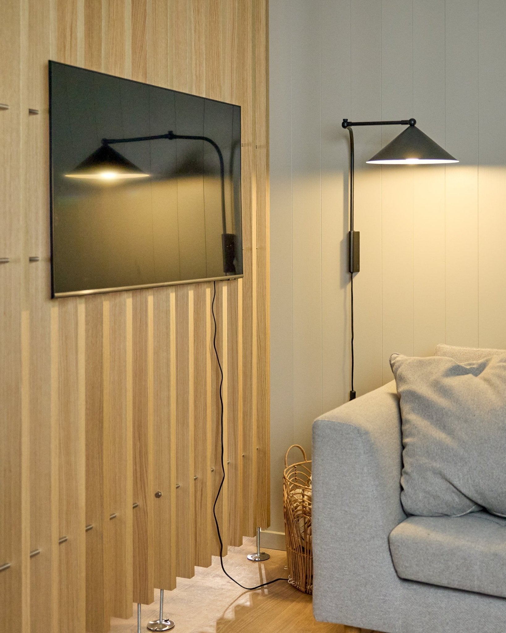 Un Nell | TV-Mount / BM1 de WoodUpp (Beige, 64x44 cm) maintient un téléviseur à écran plat sur un Panneau acoustique nordique à côté d’un canapé gris ; un panier se trouve à proximité.