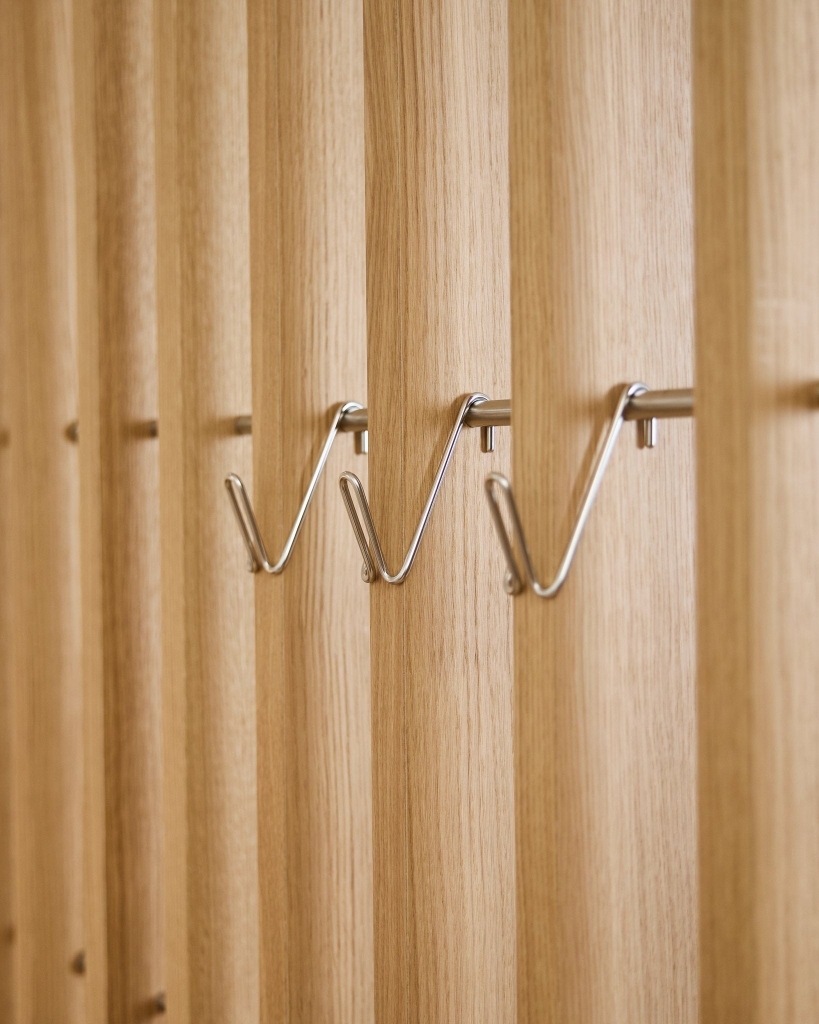 Trois accessoires Alva Wire Hook / BM1 WoodUpp, courts, en acier inoxydable steel, sont montés de façon régulière sur une même rangée sur un panneau acoustique en lattes de bois vertical.