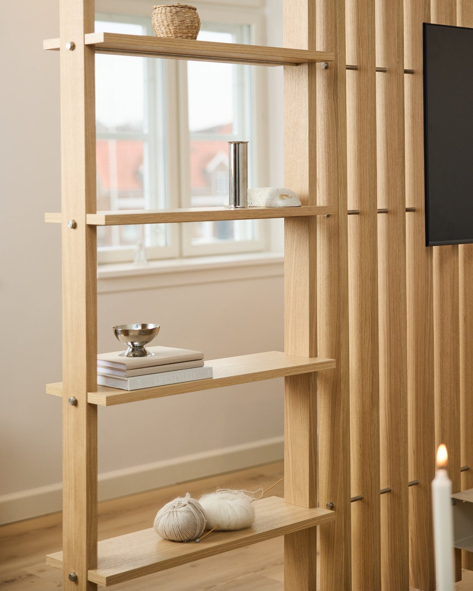 Séparateur de pièce en chêne clair signé WoodUpp, avec des lattes vertical et des étagères accueillant des livres et des pelotes de laine.