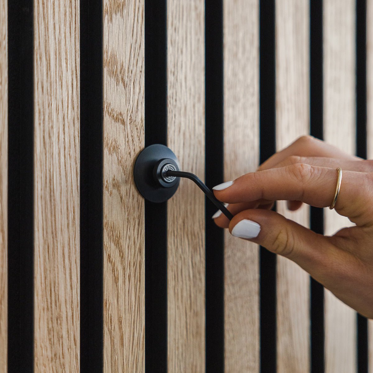 Une main aux ongles vernis de blanc tourne un crochet Dupps | Heavy en aluminium black de WoodUpp sur un panneau acoustique en bois Akupanel vertical.