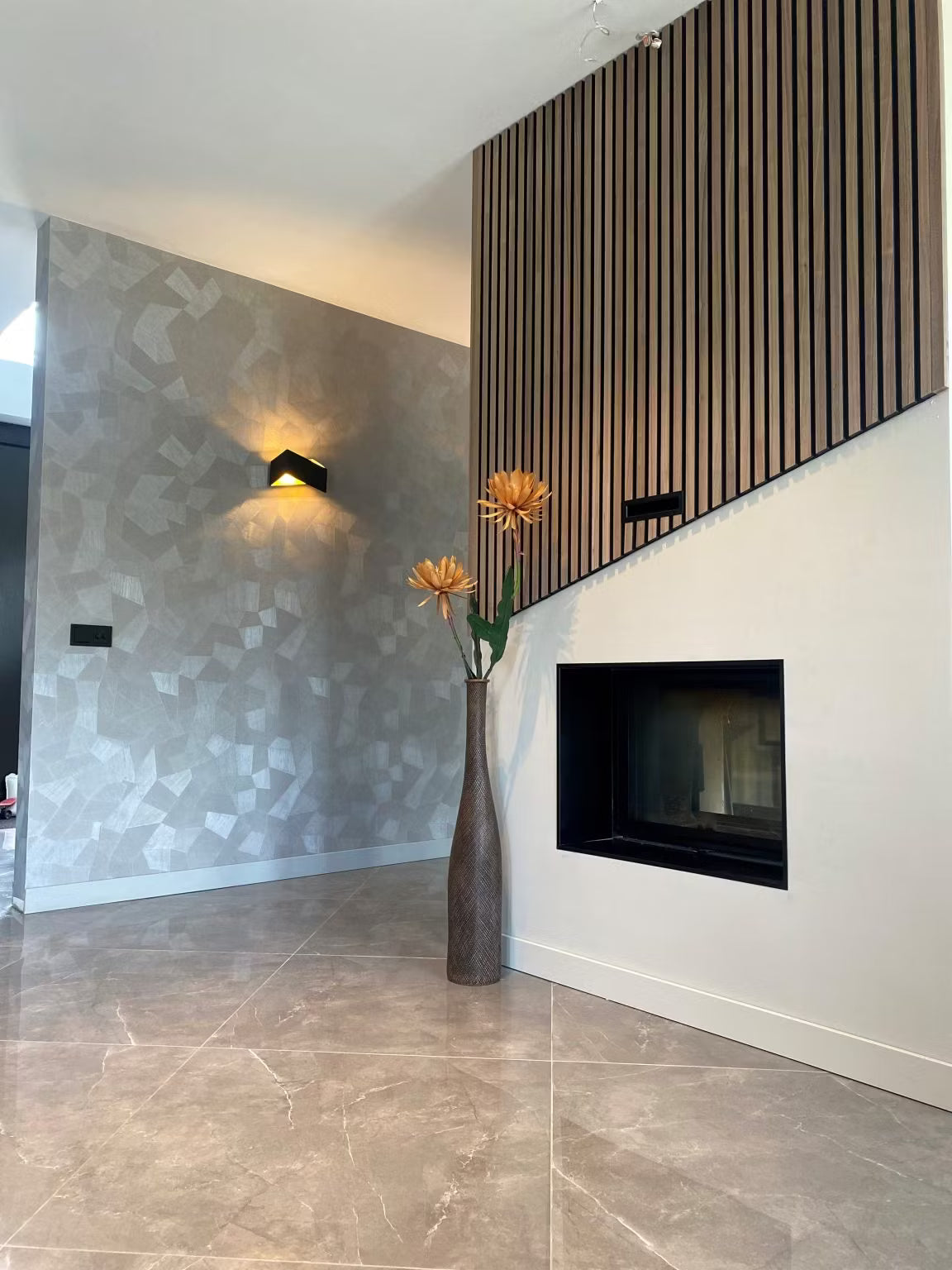 Salon moderne avec papier peint à motifs géométriques, panneaux acoustiques en bois Akupanel, cheminée murale et grand vase de fleurs orange.