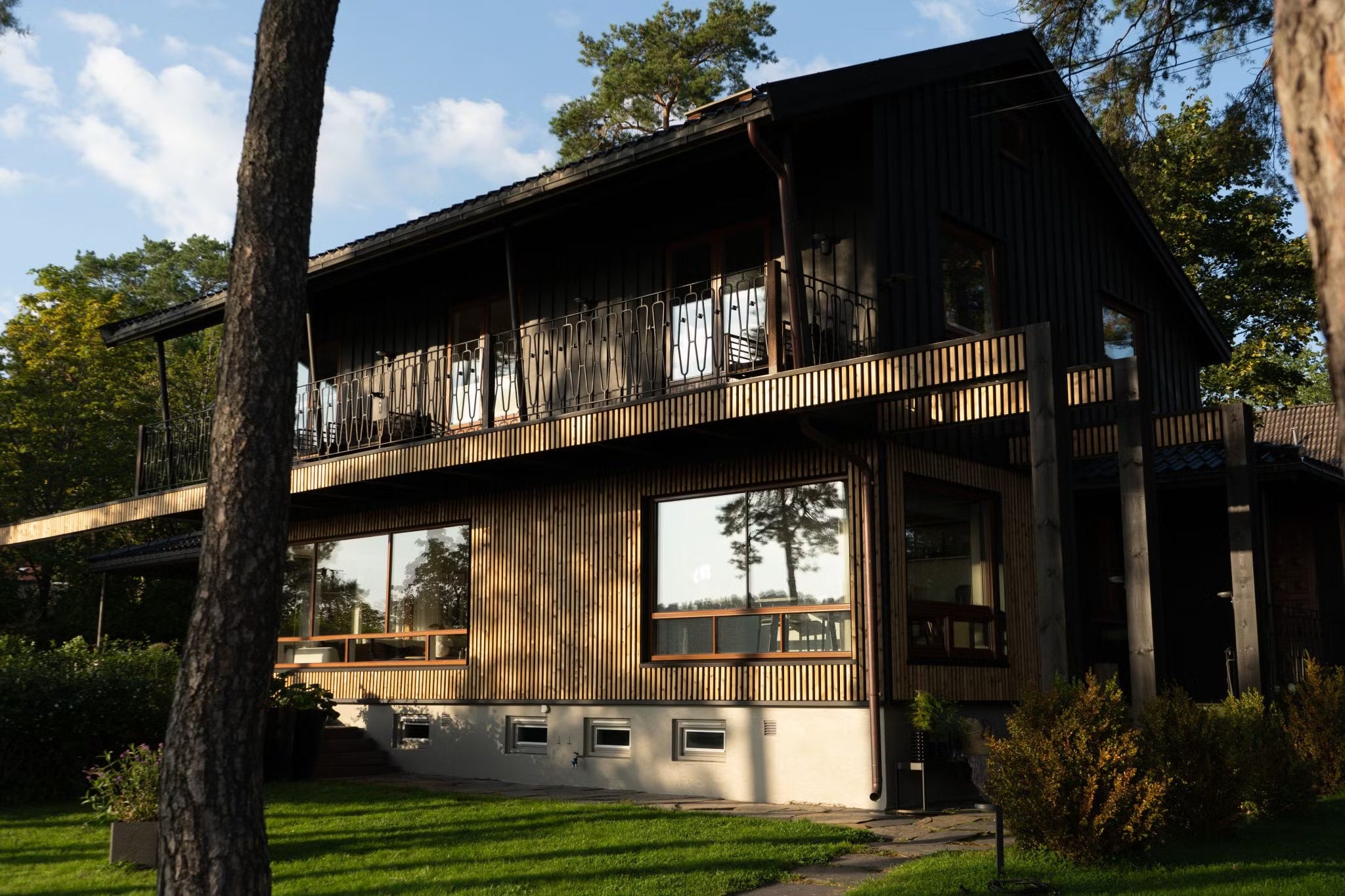 Maison moderne à deux étages avec de grandes fenêtres et des panneaux de façade en bois foncé Aluwood, entourée darbres et dune pelouse verte sous la lumière du soleil.