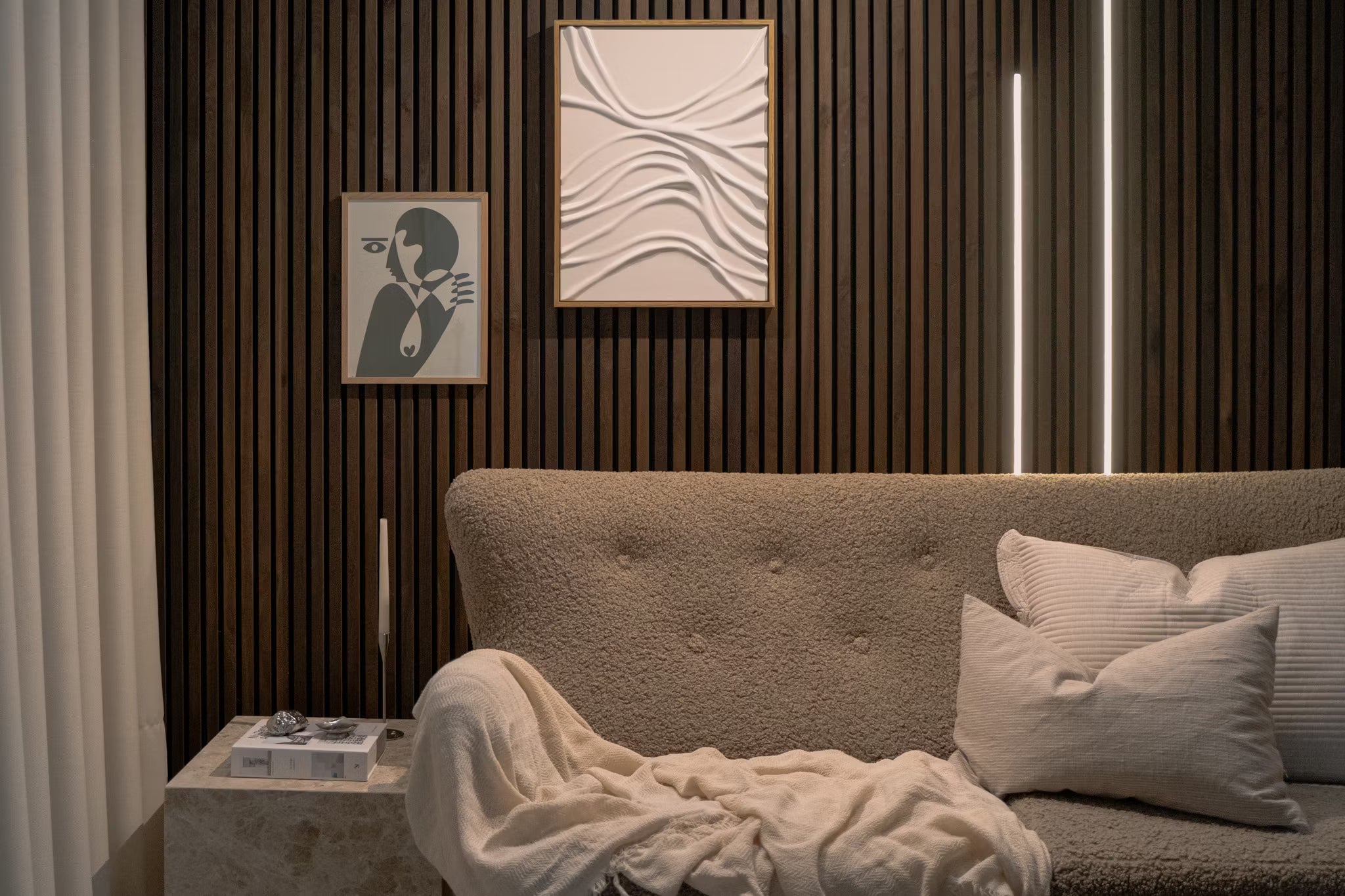 Salon moderne avec canapé beige texturé, coussins blancs, panneau acoustique abstrait et mur vertical en lattes de bois Akupanel avec un éclairage dambiance doux.