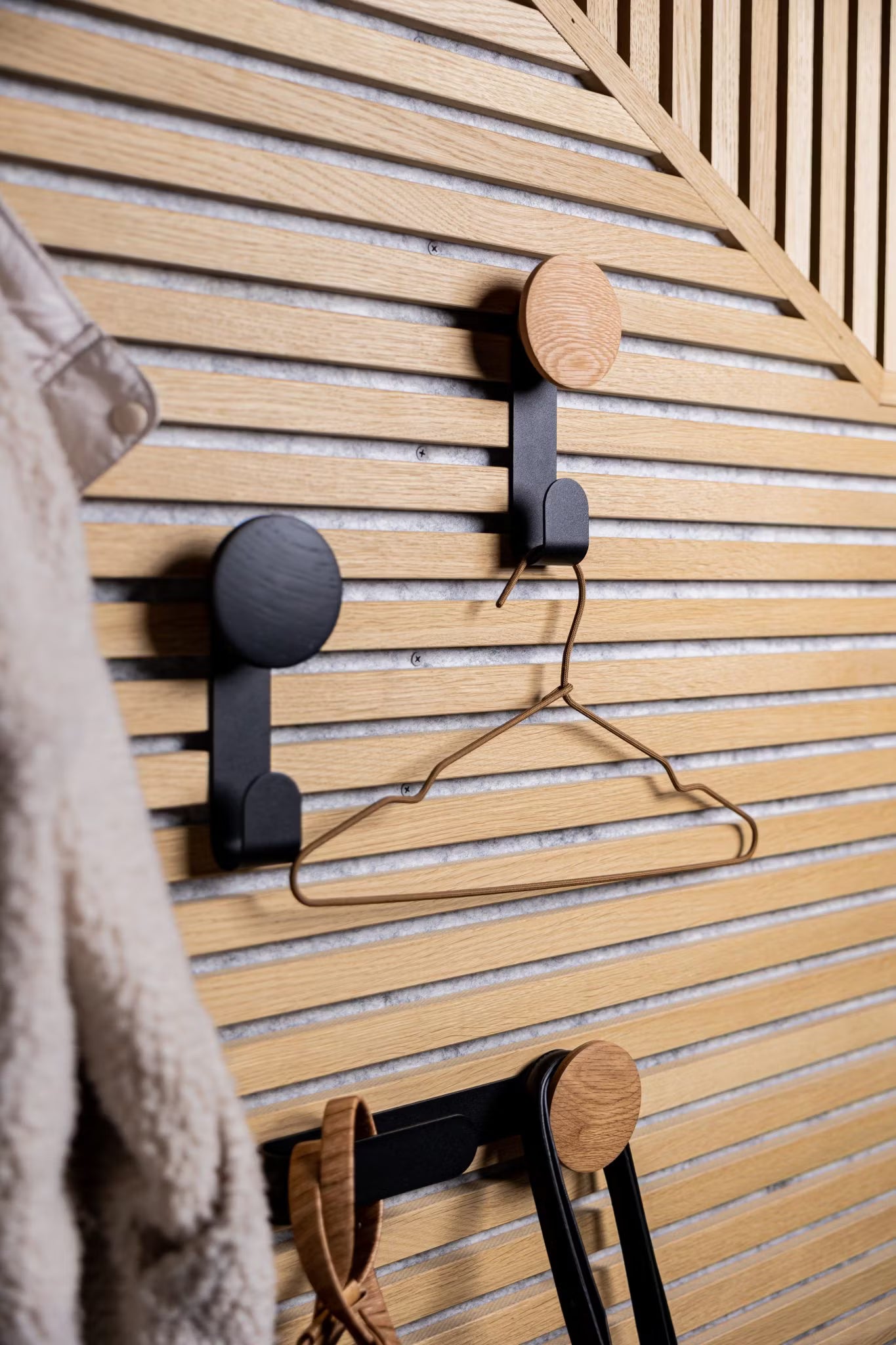 Trois crochets modernes montés sur un mur de lattes en bois acoustique Akupanel de style nordique supportent un cintre métallique et des accessoires, avec un manteau flou partiellement visible sur la gauche.