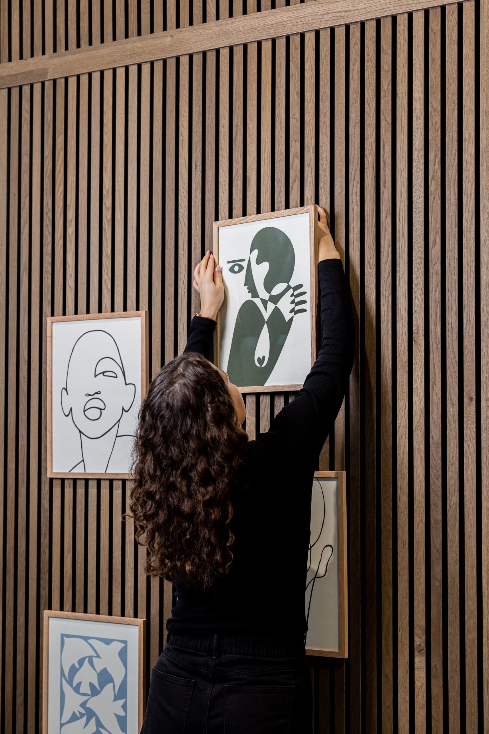 Une personne aux cheveux bouclés accroche un accessoire WoodUpp® CREATE DUPPs sur un panneau acoustique à lattes en bois, entouré d’art minimaliste et d’une décoration moderne.